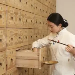 Eine junge asiatische Frau im weißen Kittel hält eine Waage in der Hand und entnimmt etwas aus einer Schublade